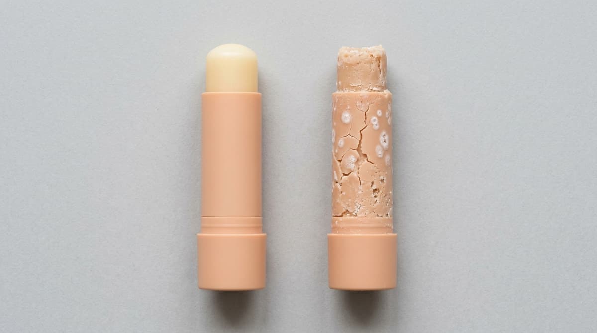 Side-by-side comparison of a perfect smooth lip balm stick and a defective one with visible surface cracks, white bloom spots and uneven texture, flat lay on light grey background, educational cosmetic chemistry photography